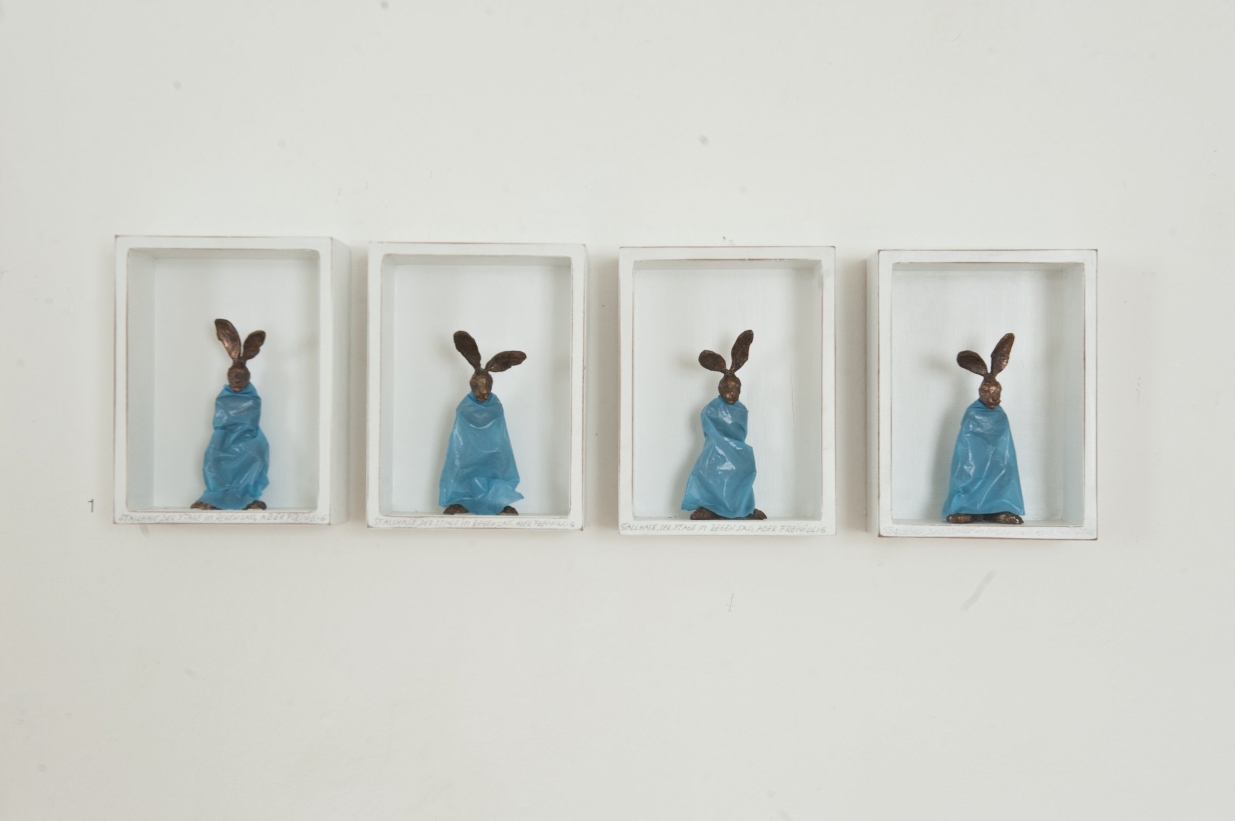 Kleine Hasenskulpturen mit blauen Regenumhängen (Mülltüten) in weißen, eckigen Bilderrahmen